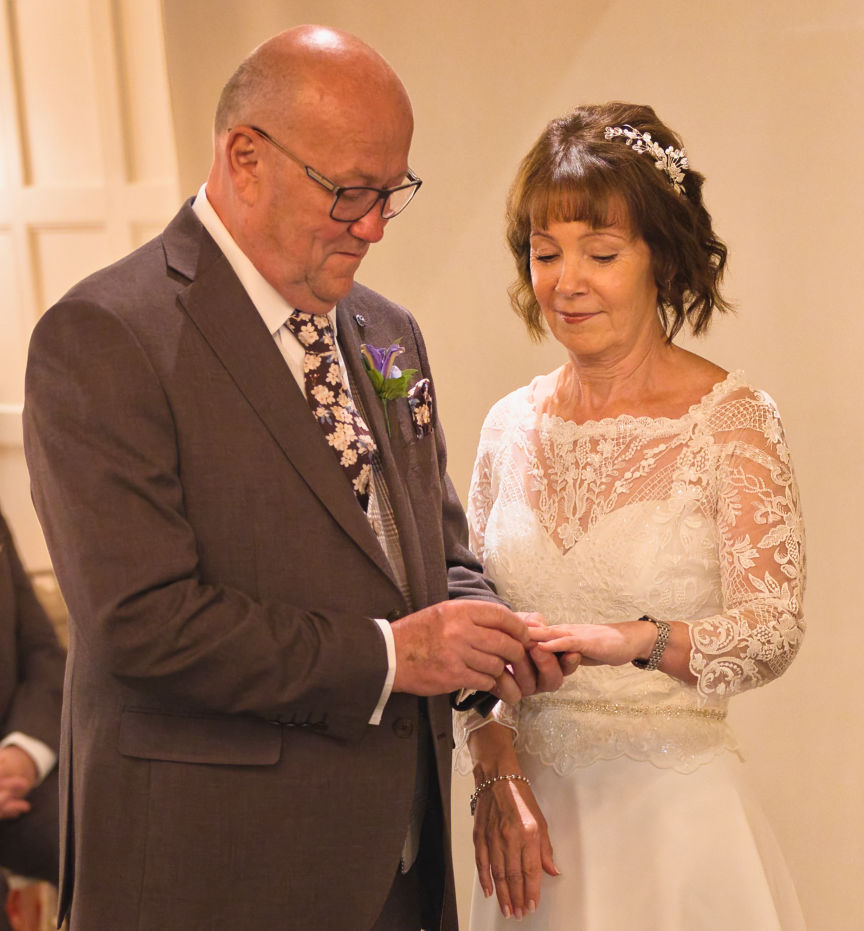 A mature groom placing the ring on his bride's finger in the grill room inside Stanneylands Hotel Winslow